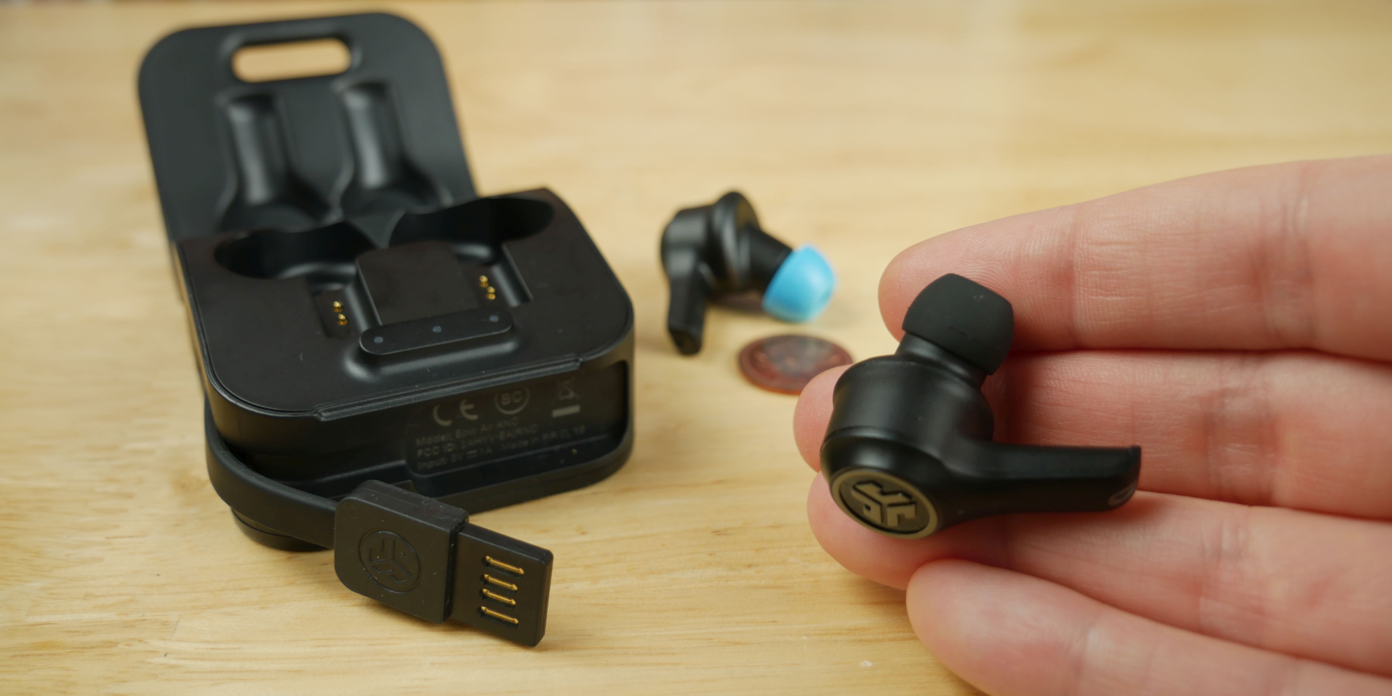 Holding an Epic Air ANC earbud in one hand with charging case and other earbud in the background.