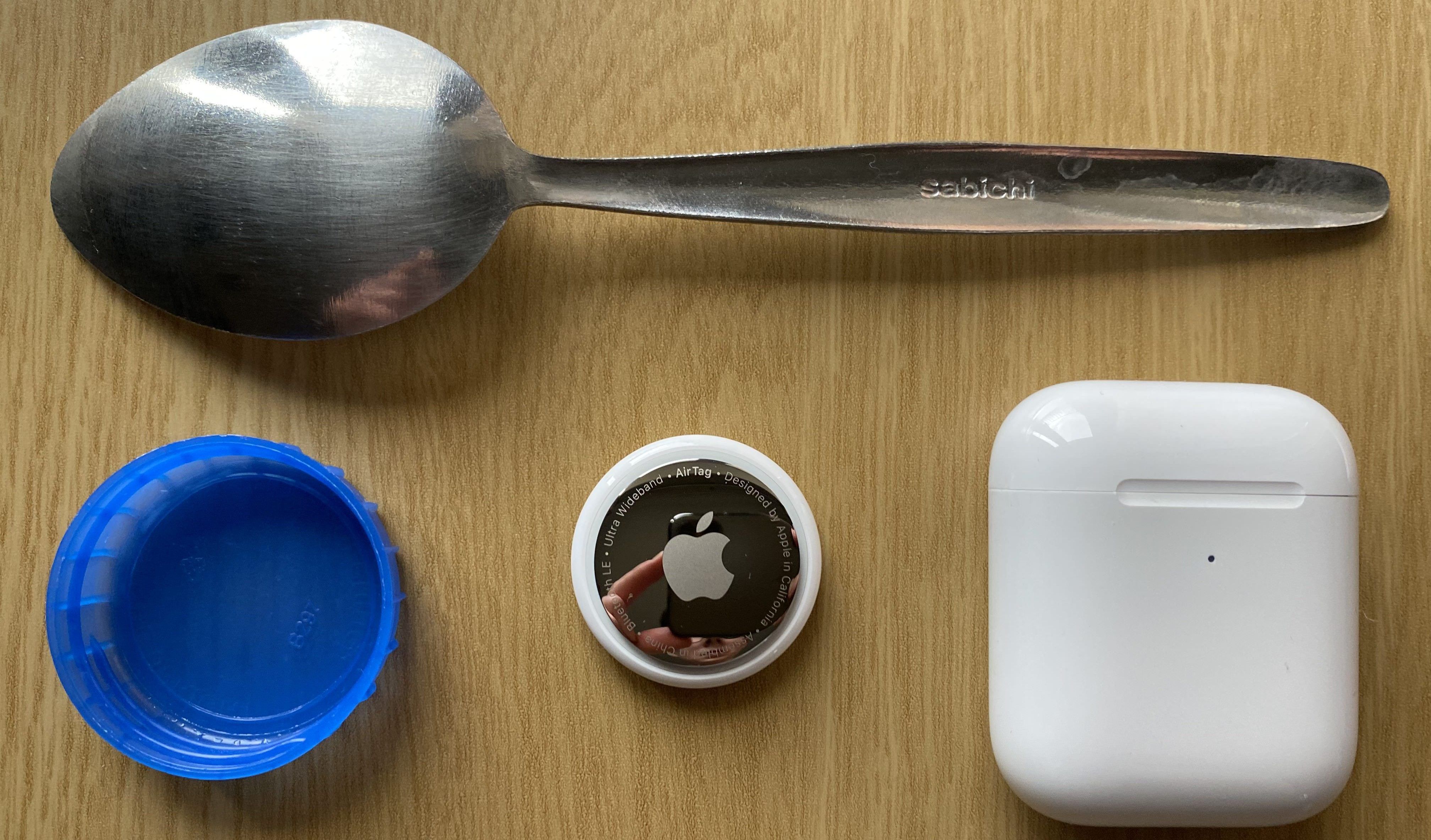 An AirTag next to a milk bottle cap, a spoon, and an AirPods case to show the size