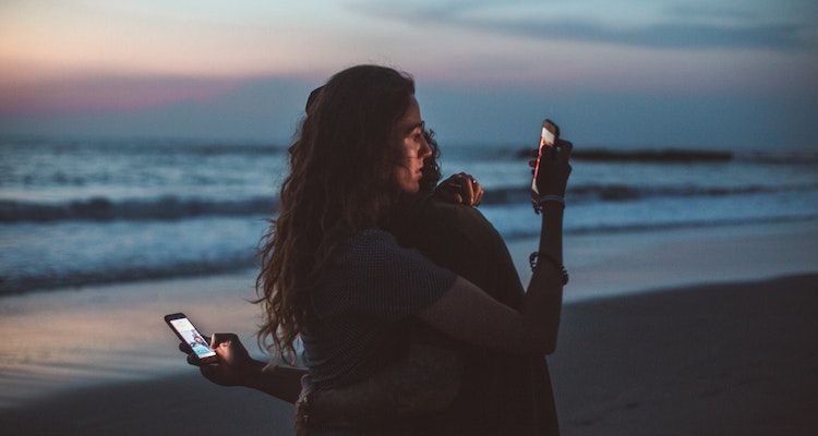 A couple hugging and looking at their phones on the beach