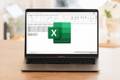 A Laptop Sitting On Top of a Wooden Table. An Excel Spreadsheet Is Blurred Out in the Background, and the Excel Logo is in the Foreground