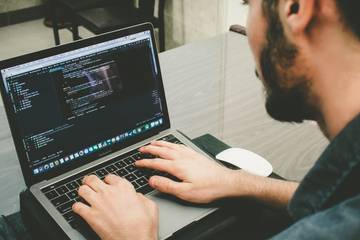 A man sitting at a desk typing on a laptop with code on the screen.