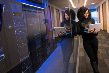 A woman stands next to a server rack holding a laptop.
