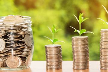A jar filled with coins and three stacks of coins with saplings growing out of the top of each.