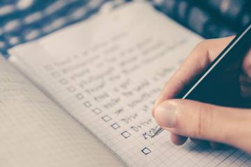 A hand holding a pen, hovering over a list of items handwritten on paper.