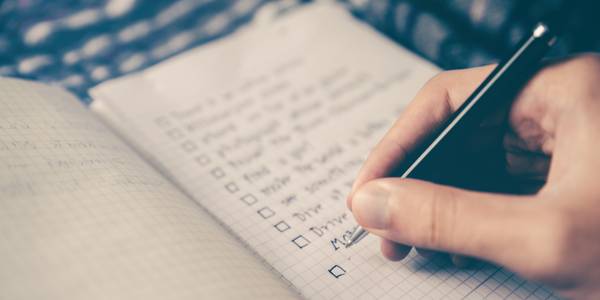 A hand holding a pen, hovering over a list of items handwritten on paper. 