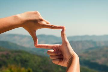 Two hands held with thumbs and forefingers extended to form a rectangle in the central foreground of a landscape scene