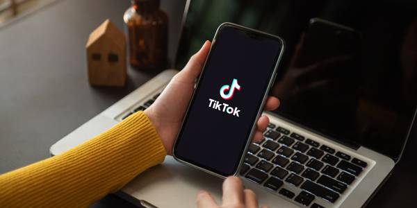 a hand holding a smartphone with the tiktok app logo and a laptop in the background