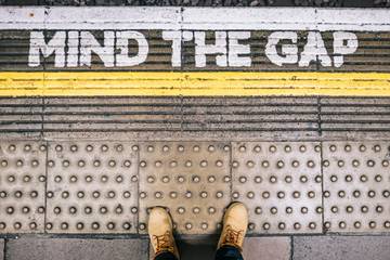 A pair of feet seen from above, near the edge of a station platform with “Mind The Gap” written on the floor