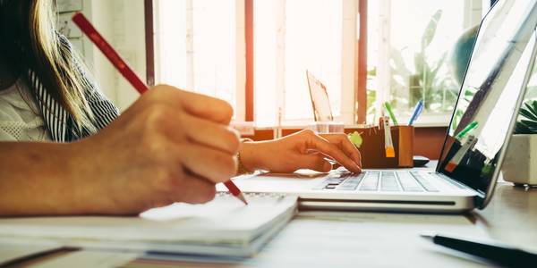 A close-up of a hand writing with a pencil in front of another hand typing on a laptop