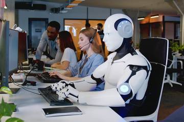 A robot sits at a desk appearing to type on a keyboard, working alongside human employees.