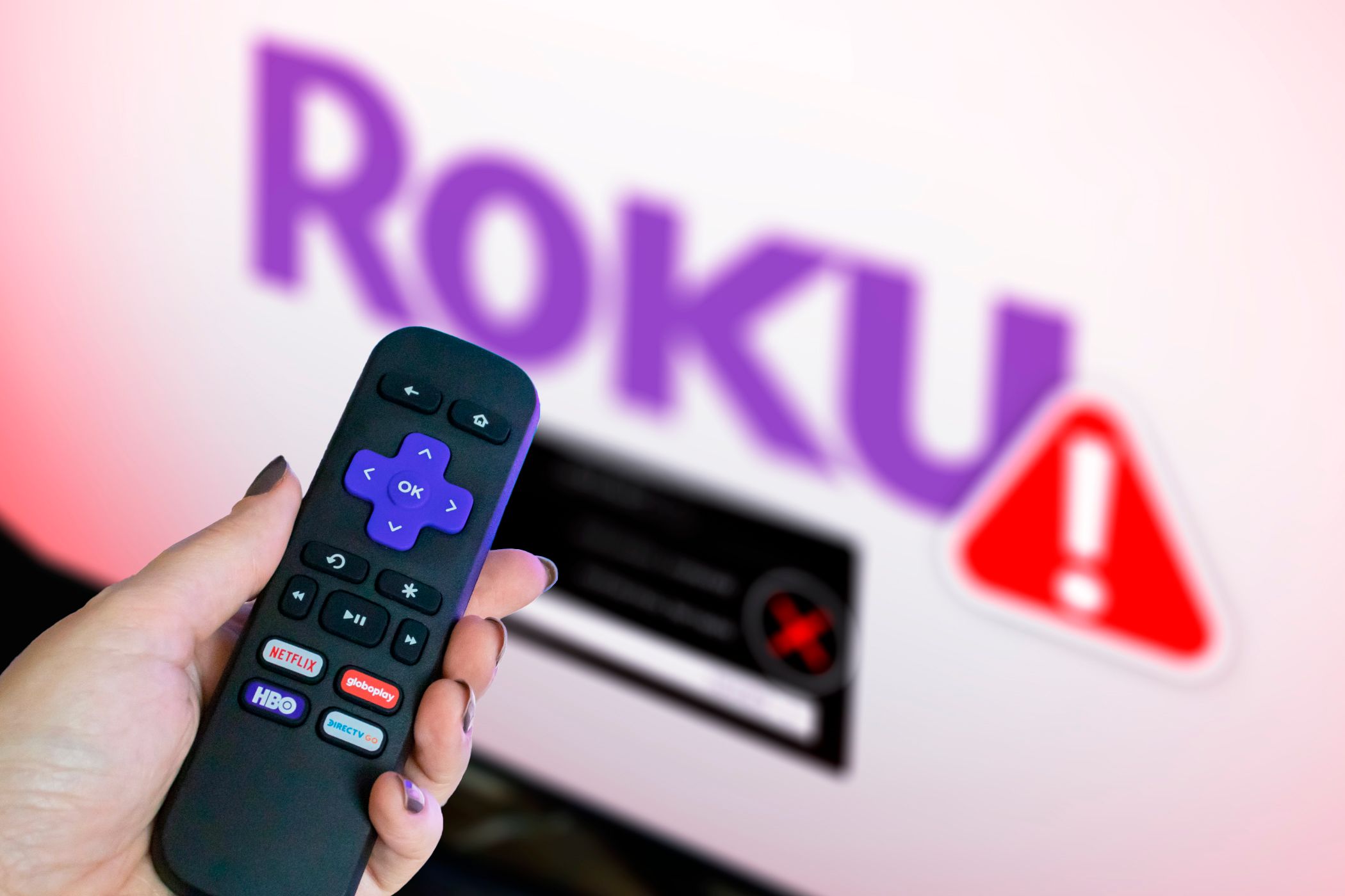 A hand holding a Roku remote with a TV in the background showing the Roku logo blurred and an alert.