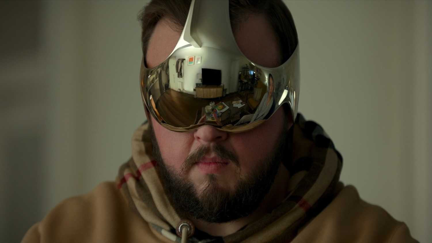 A man in a hoodie wears a reflective metallic visor showing a living room and another person, from Netflix’s 3 Body Problem