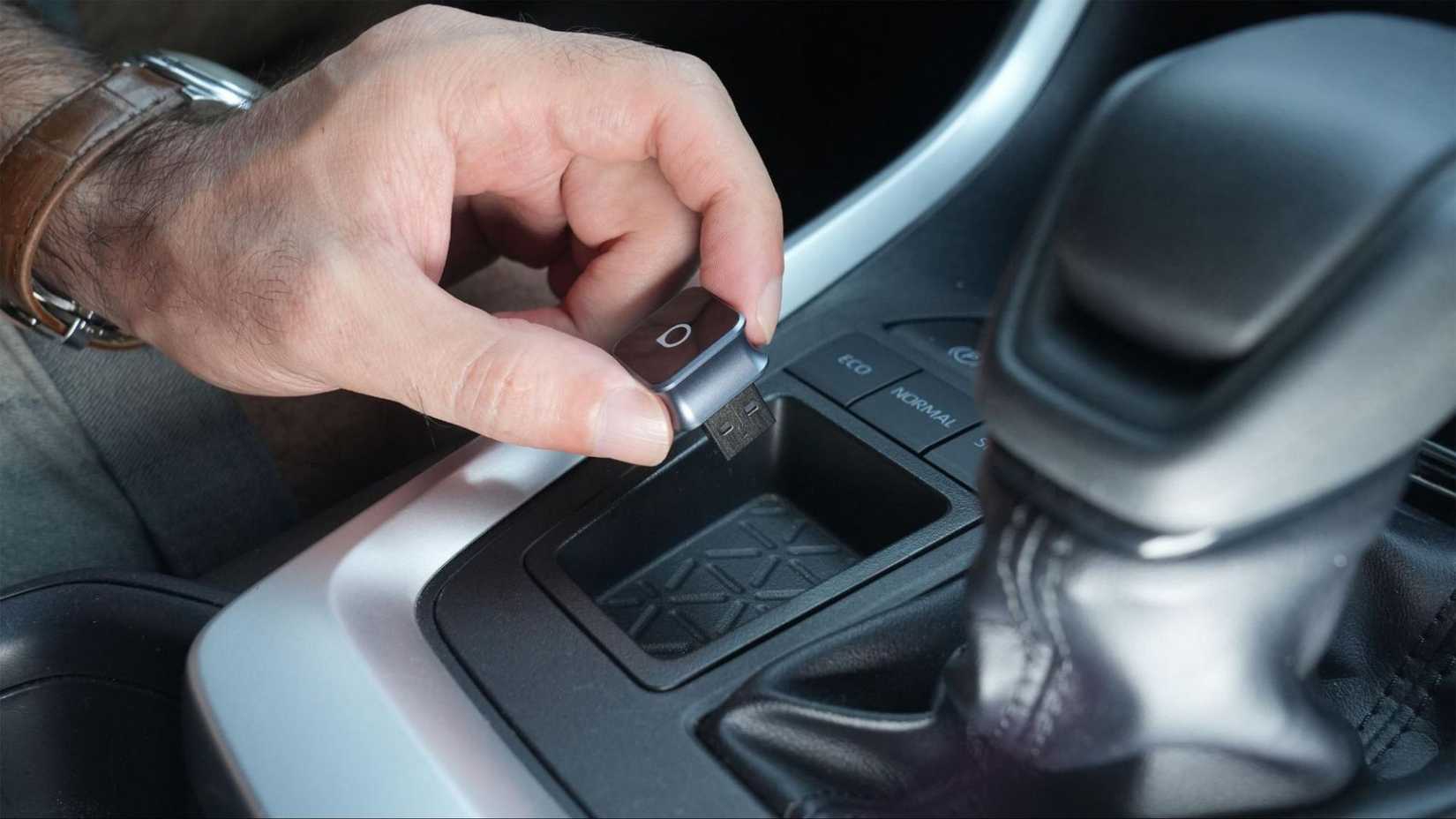 A man's hand holds an Ottocast MINI Cube next to a gear lever.