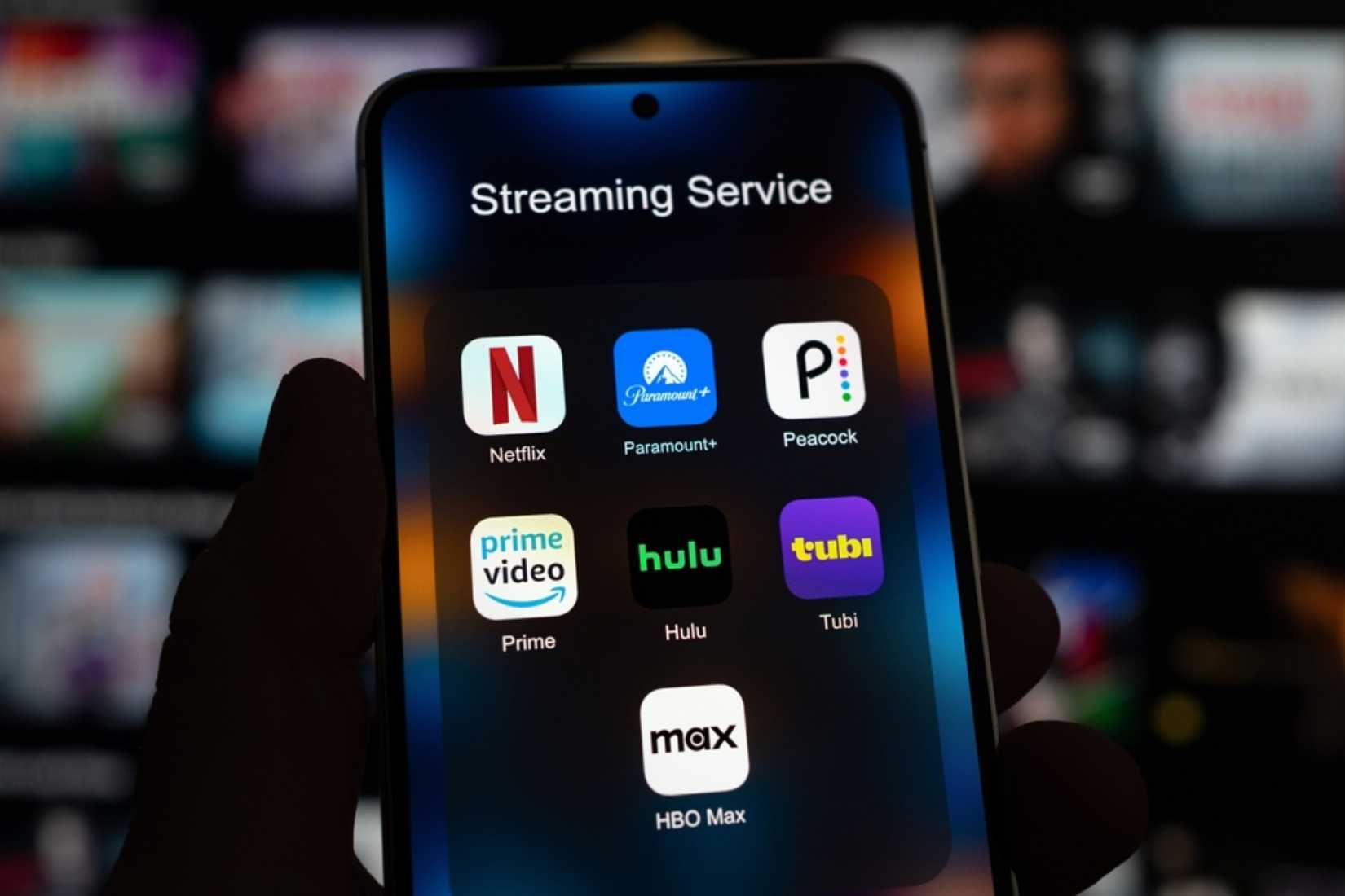 Person holding a phone showing streaming apps including Netflix, Paramount, Peacock, Prime, Hulu, Tubi, HBO Max