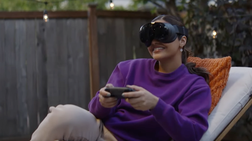 A person wears the Valve Steam Frame and uses the controllers to navigate a virtual experience. The person is sitting on a couch outside.
