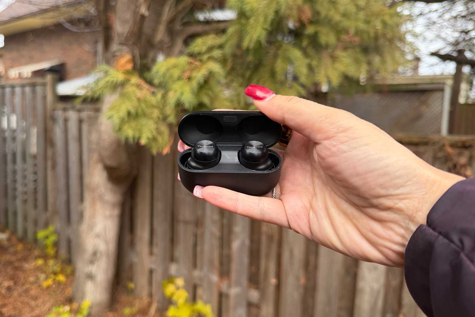 The Panasonic Technics EAH-AZ100 earbuds in hand in their case outside with trees in the background.