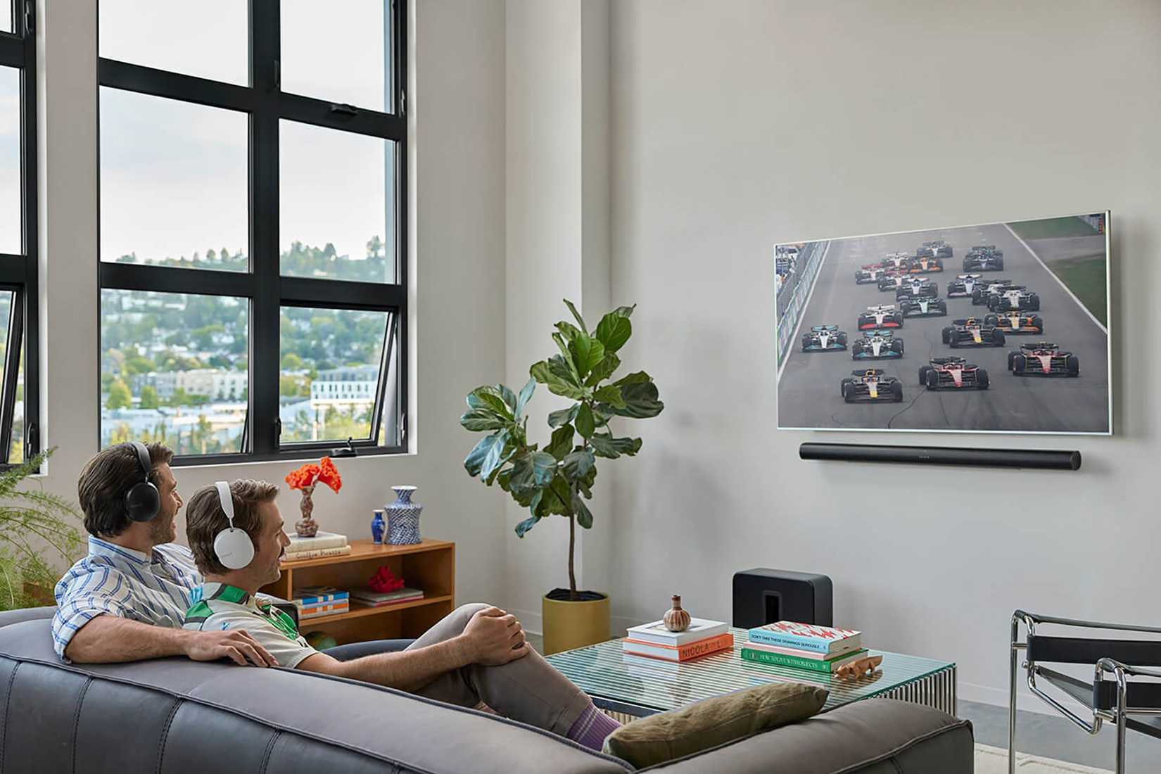 Two men on a couch watching TV, both wearing Sonos Ace headphones.