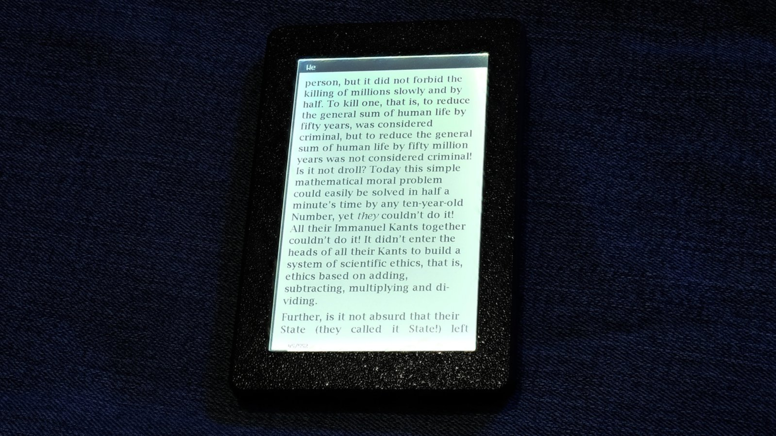 A close-up of the Open Book Touch e-reader displaying a page of text from an e-book in a textured, black snap-fit enclosure against a dark blue fabric background.