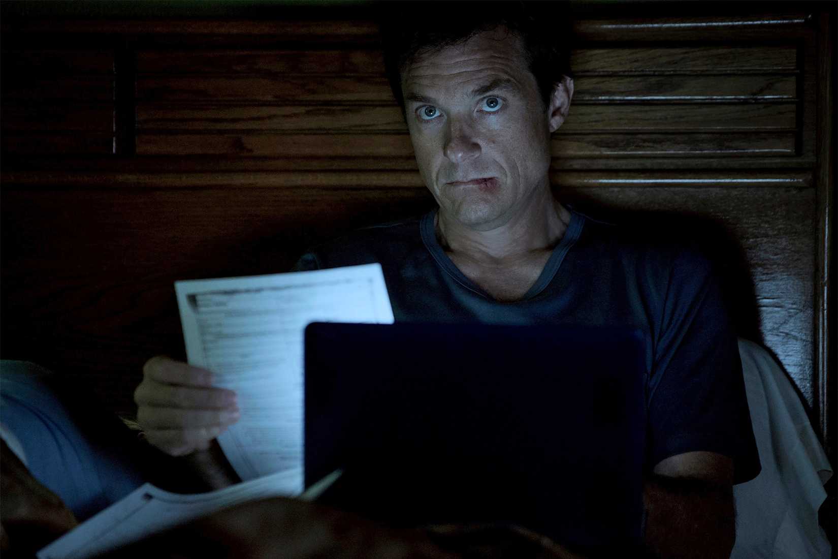 Marty lying in bed with a busted lip looking at papers and his computer in Ozark.