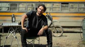 Link sitting on a chair outside in front of a bus, his hand on his thigh in Paradise.