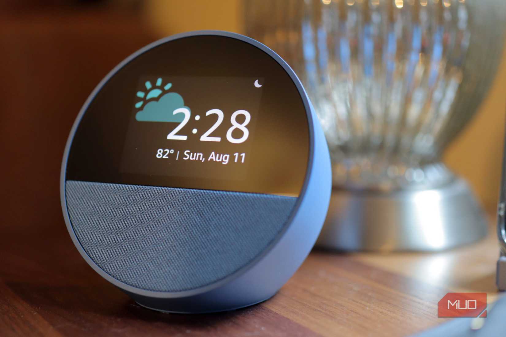 Blue Amazon Echo Spot on a wooden surface with a silver lamp in the background.