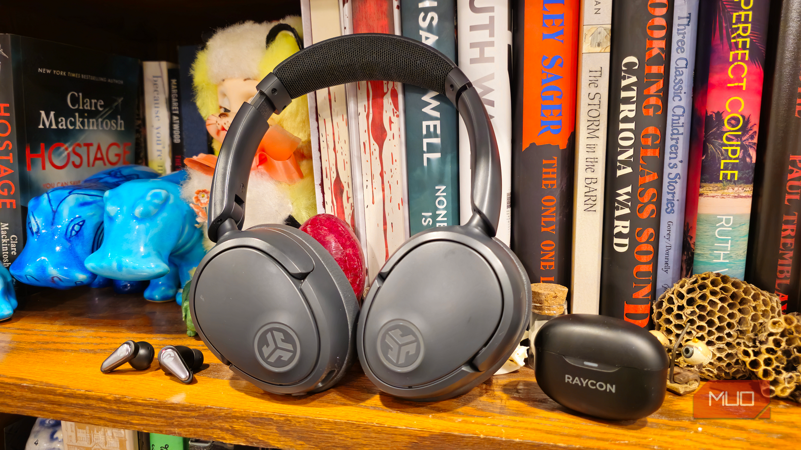 A pair of Raycon Everyday Pro Earbuds and JBuds Lux ANC Over-Ear Headphones on a bookcase