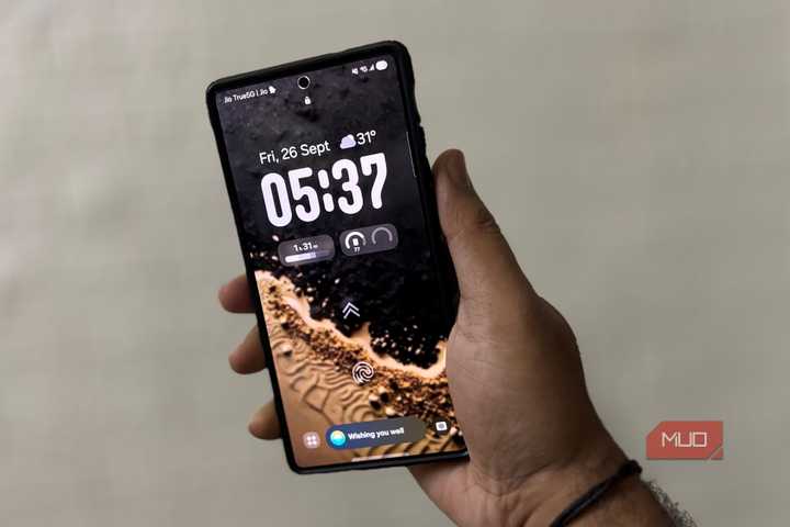Hand holding a Samsung Galaxy with One UI lock screen, showing date, time, and widgets.