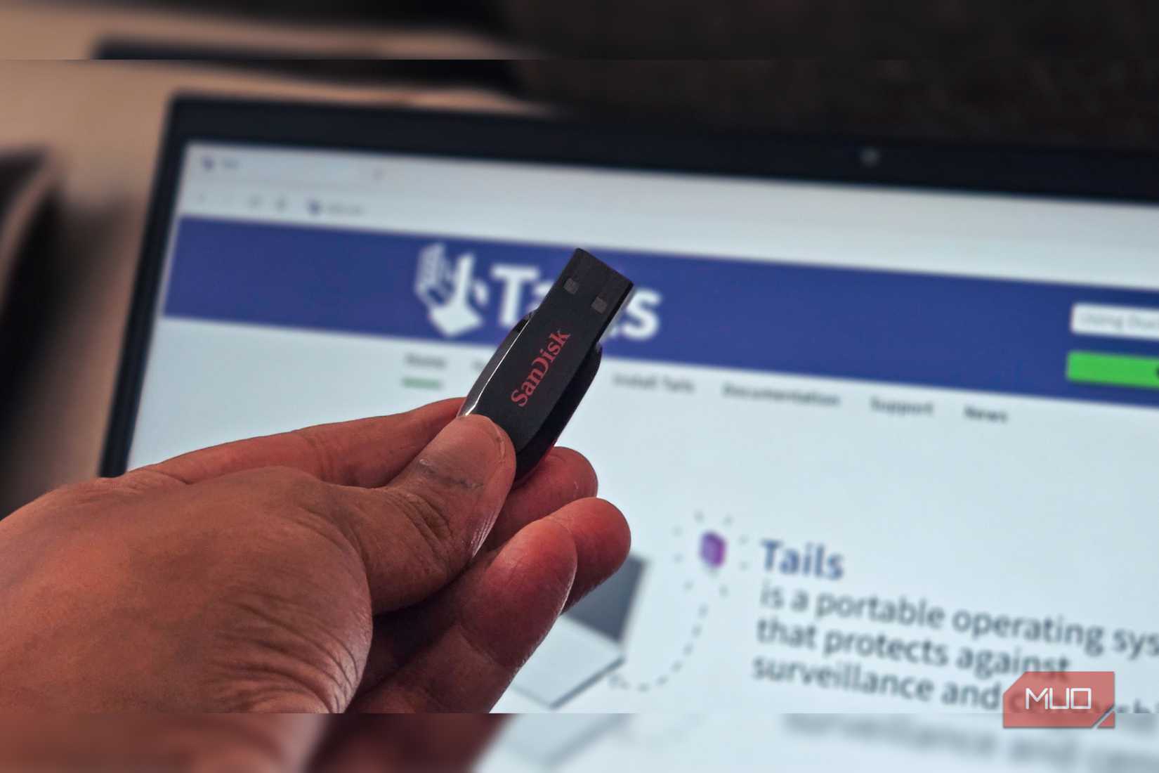 A personal hand holding a Sandisk USB flash drive and a laptop in the background