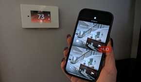 A hand holding a phone by a smart thermostat showing a smart home app.