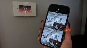 A hand holding a phone by a smart thermostat showing a smart home app.
