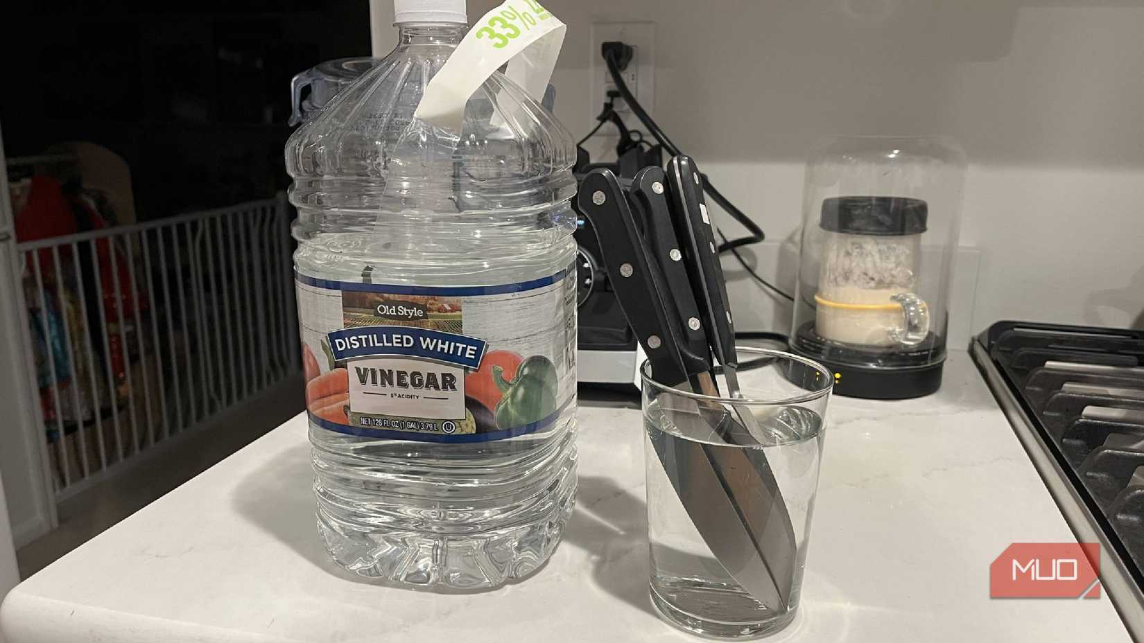 A jug of white vinegar next to knives in a cup of white vinegar