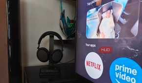 The Sennheiser RS 275 headphones on a stand with the BTA1 Bluetooth transmitter at the base beside a TV.