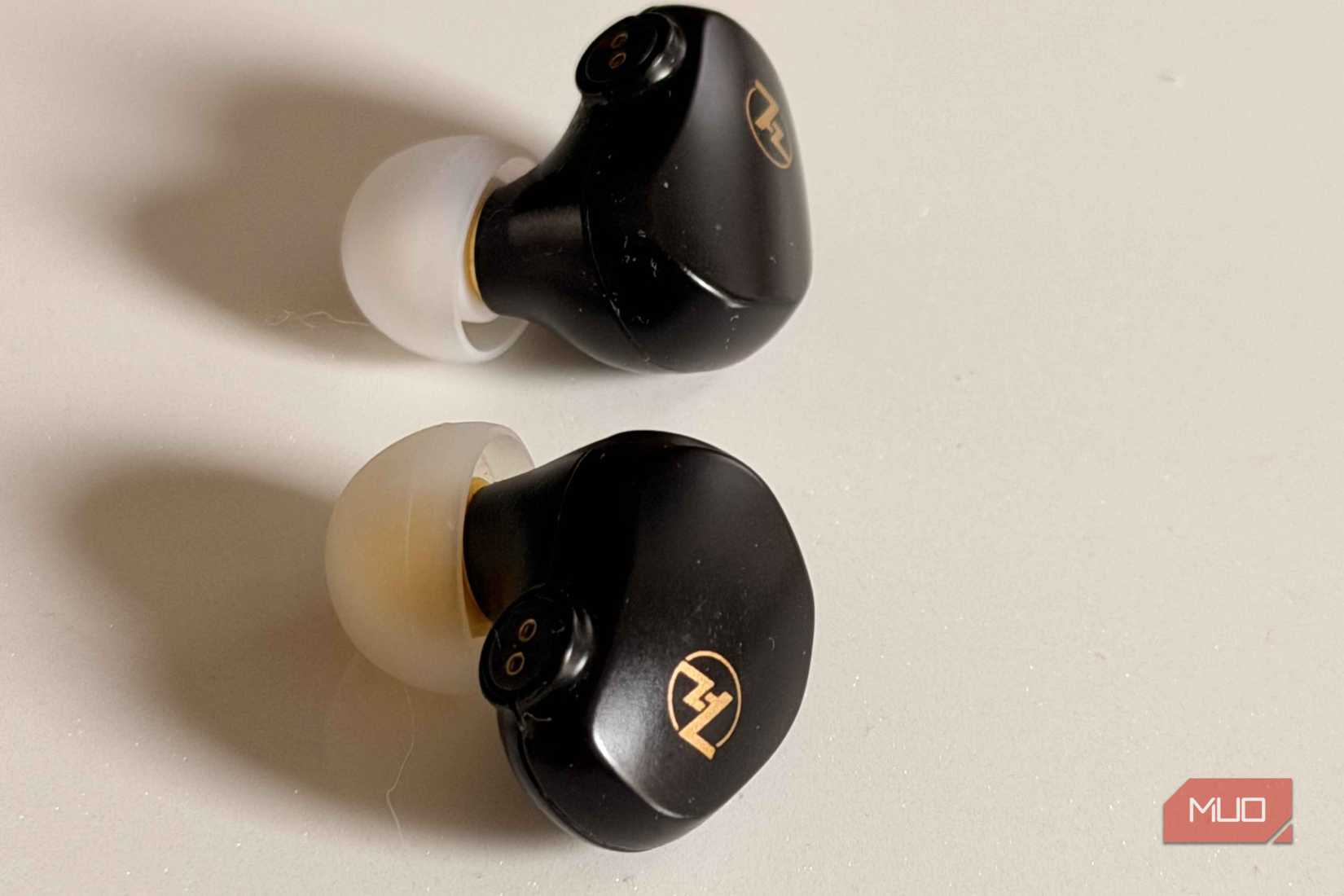 Close up of two black IEM earphones showing mmcx connectors and gold logos with silicone tips on a beige surface