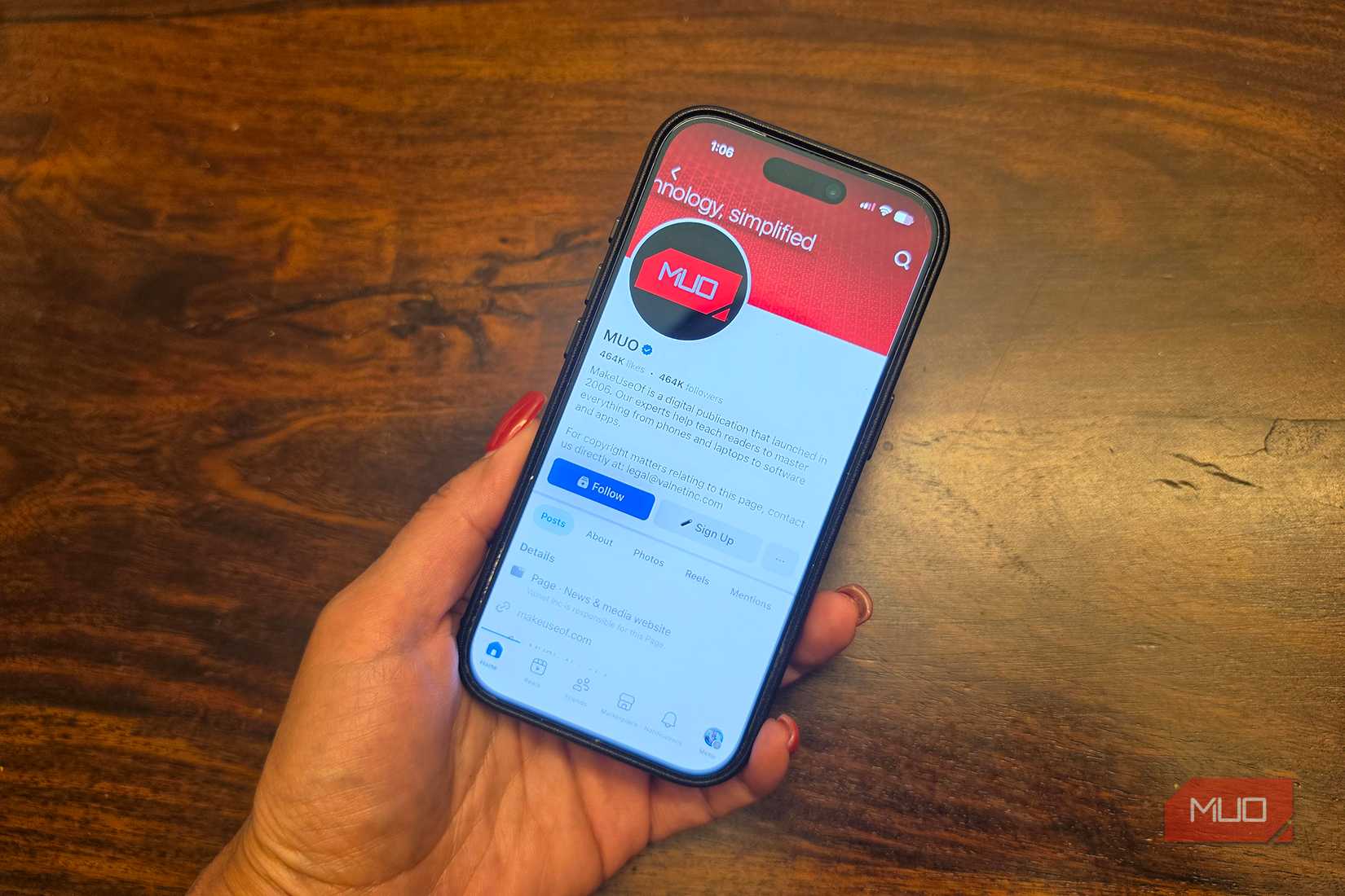 A hand holding an iPhone showing the MUO Facebook page.