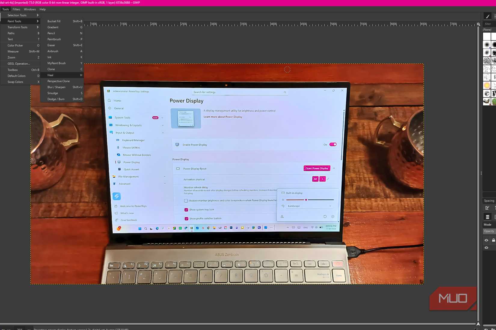 GIMP showing Tools > Paint Tools > Heal over a photo of a laptop on a wooden table, with the Heal brush positioned over the wood