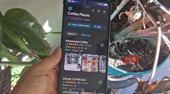A hand holding a smartphone showing a Google Maps Coffee Places list beside several potted plants